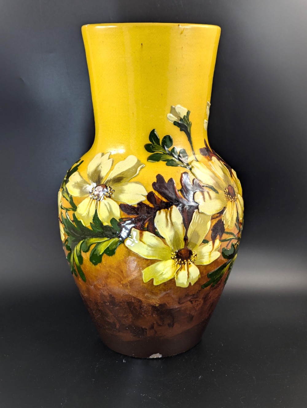 A tall ceramic vase featuring a yellow neck and upper body with raised impasto floral decoration transitioning into a dark brown glazed base. View II