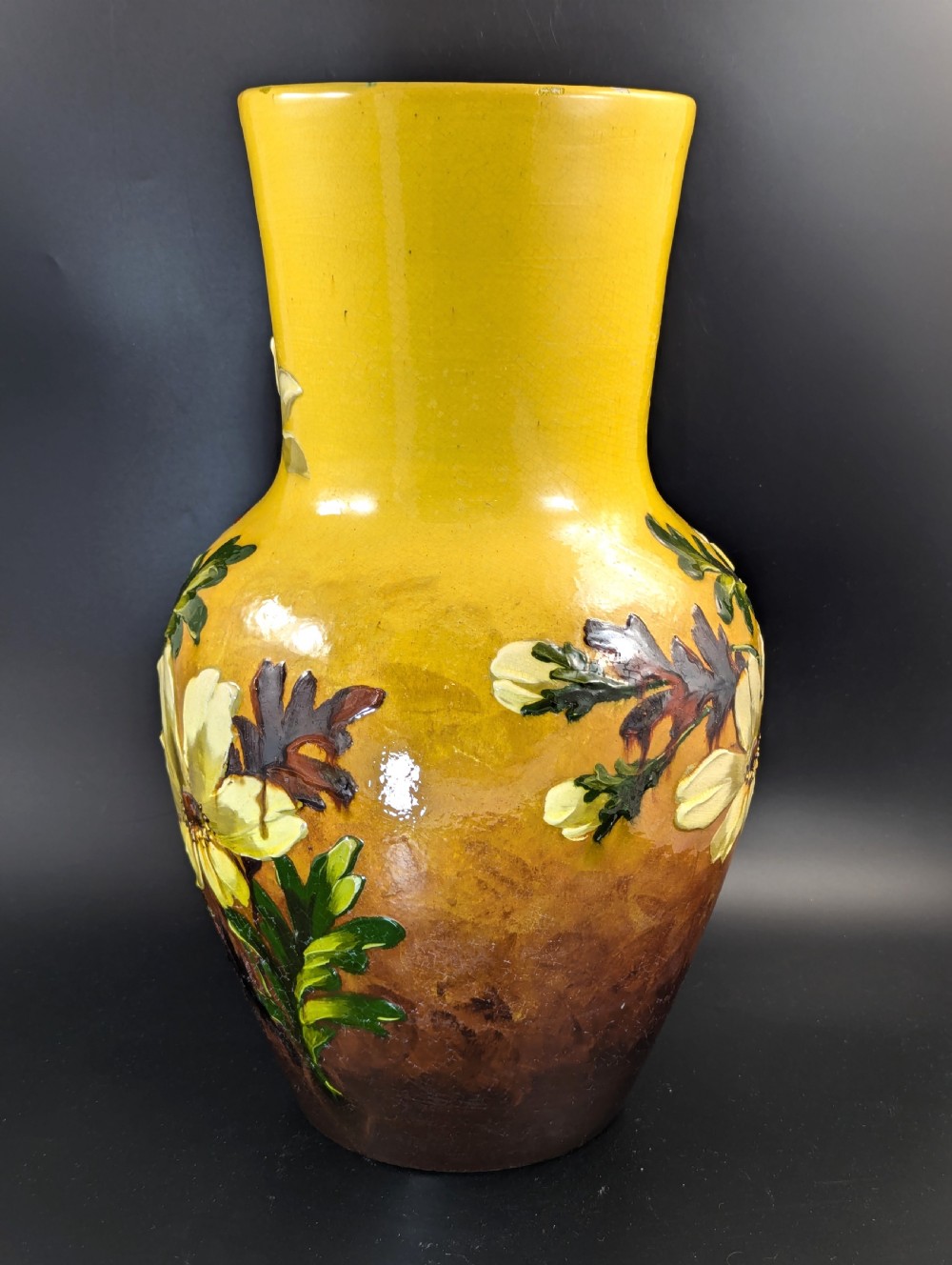A tall ceramic vase featuring a yellow neck and upper body with raised impasto floral decoration transitioning into a dark brown glazed base. View V