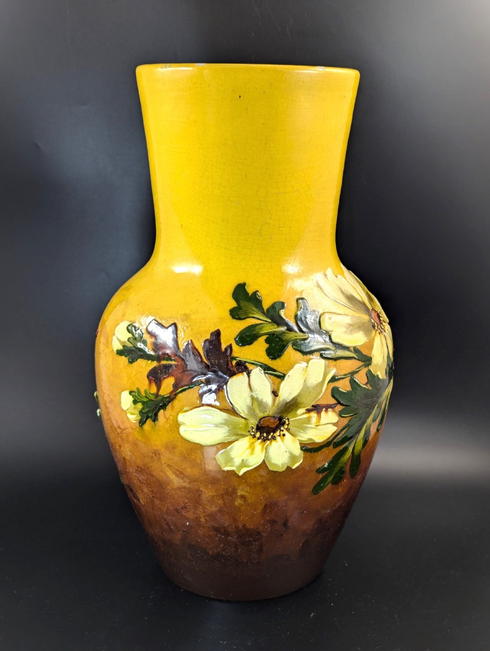 A tall ceramic vase featuring a yellow neck and upper body with raised impasto floral decoration transitioning into a dark brown glazed base. View VI