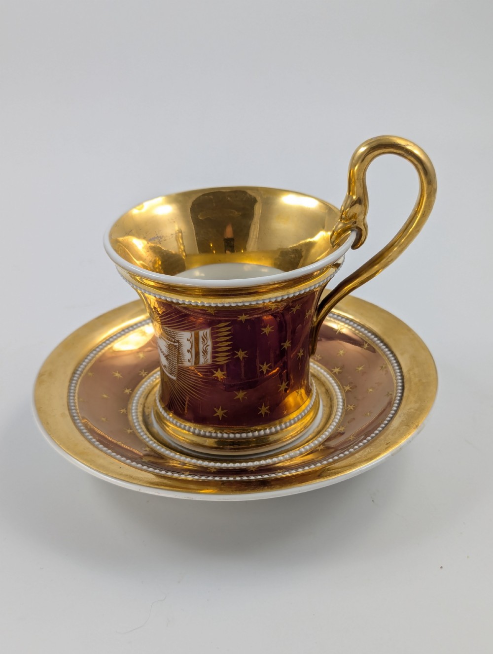 A Neoclassical KPM Berlin porcelain teacup and saucer featuring heavy gold leaf gilding, a maroon background with star patterns, and a high curved loop handle.