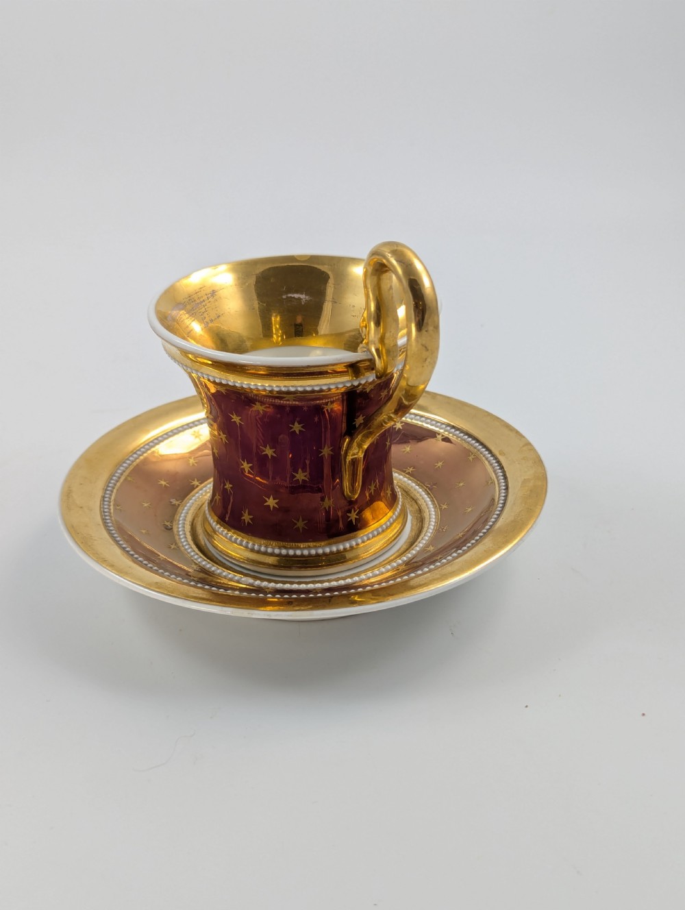 A Neoclassical KPM Berlin porcelain teacup and saucer featuring heavy gold leaf gilding, a maroon background with star patterns, and a high curved loop handle. View II