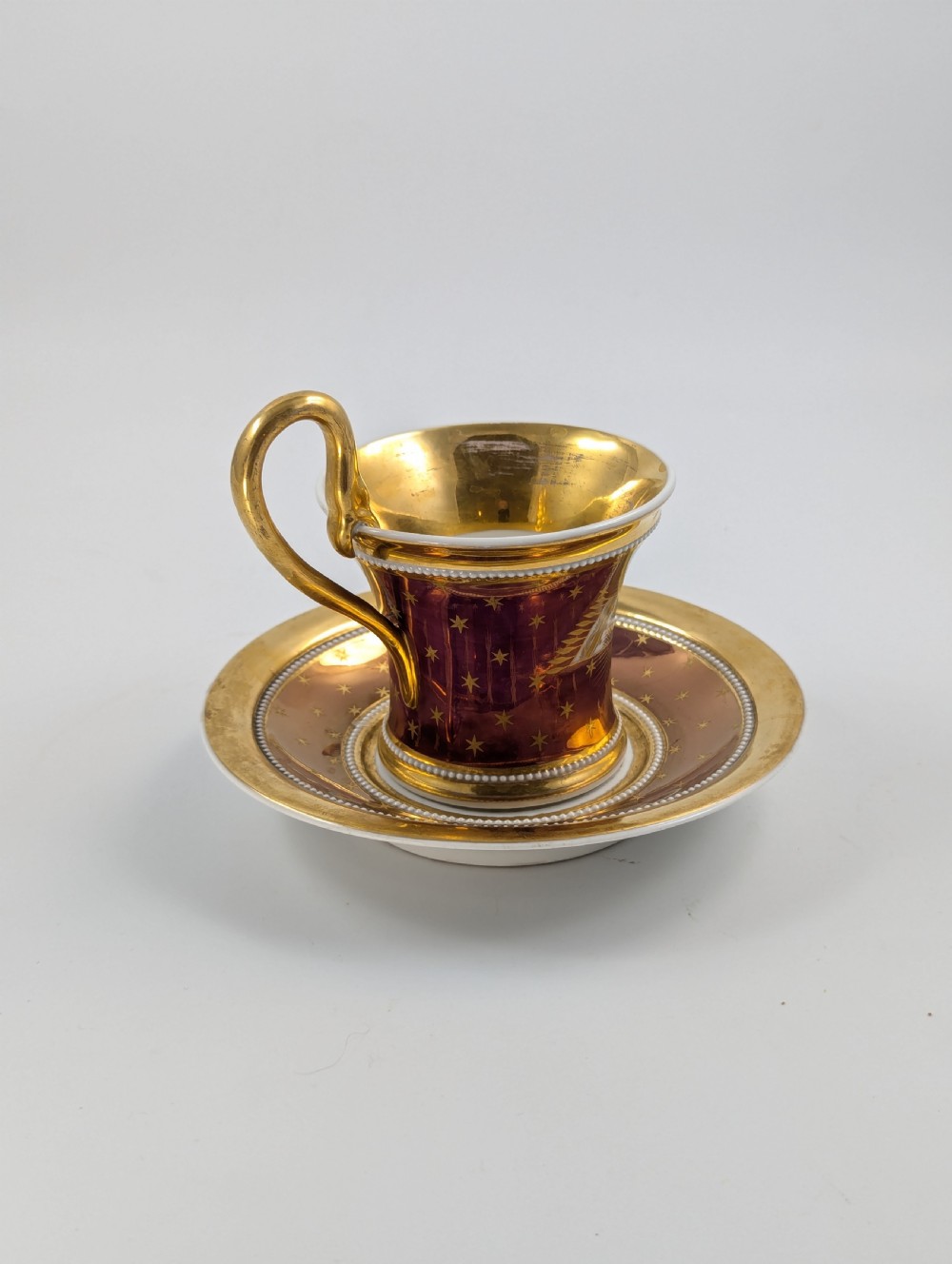 A Neoclassical KPM Berlin porcelain teacup and saucer featuring heavy gold leaf gilding, a maroon background with star patterns, and a high curved loop handle. View III