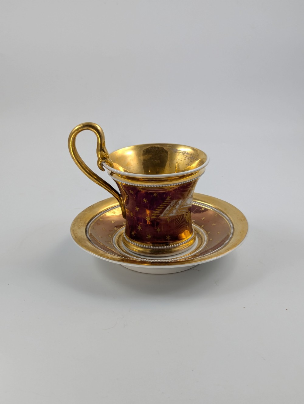 A Neoclassical KPM Berlin porcelain teacup and saucer featuring heavy gold leaf gilding, a maroon background with star patterns, and a high curved loop handle. View IV