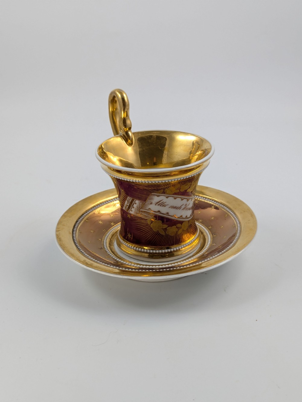 A Neoclassical KPM Berlin porcelain teacup and saucer featuring heavy gold leaf gilding, a maroon background with star patterns, and a high curved loop handle. View V