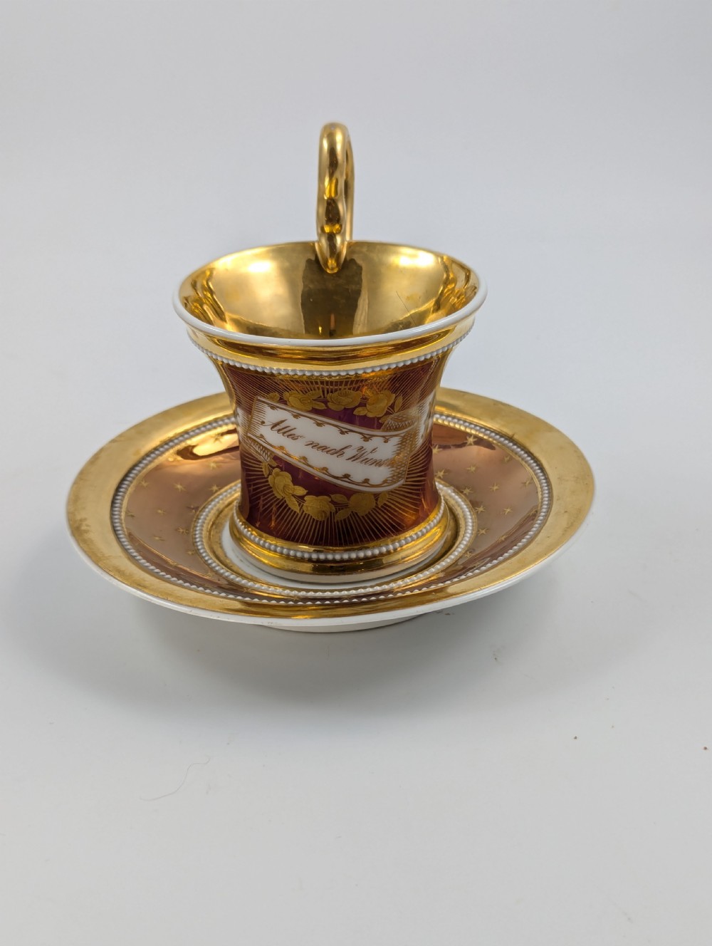 A Neoclassical KPM Berlin porcelain teacup and saucer featuring heavy gold leaf gilding, a maroon background with star patterns, and a high curved loop handle. View VI