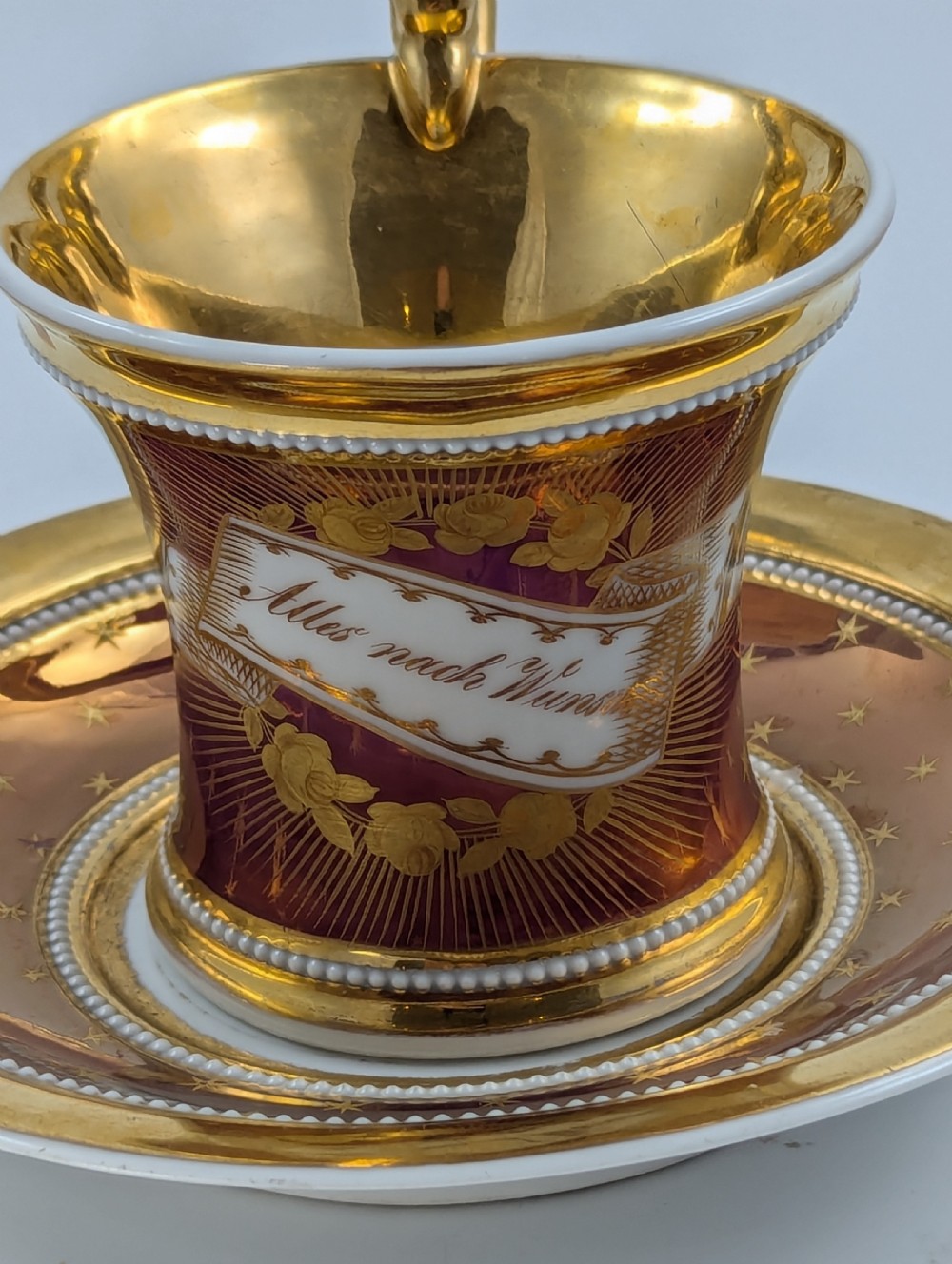 A Neoclassical KPM Berlin porcelain teacup and saucer featuring heavy gold leaf gilding, a maroon background with star patterns, and a high curved loop handle. View VII