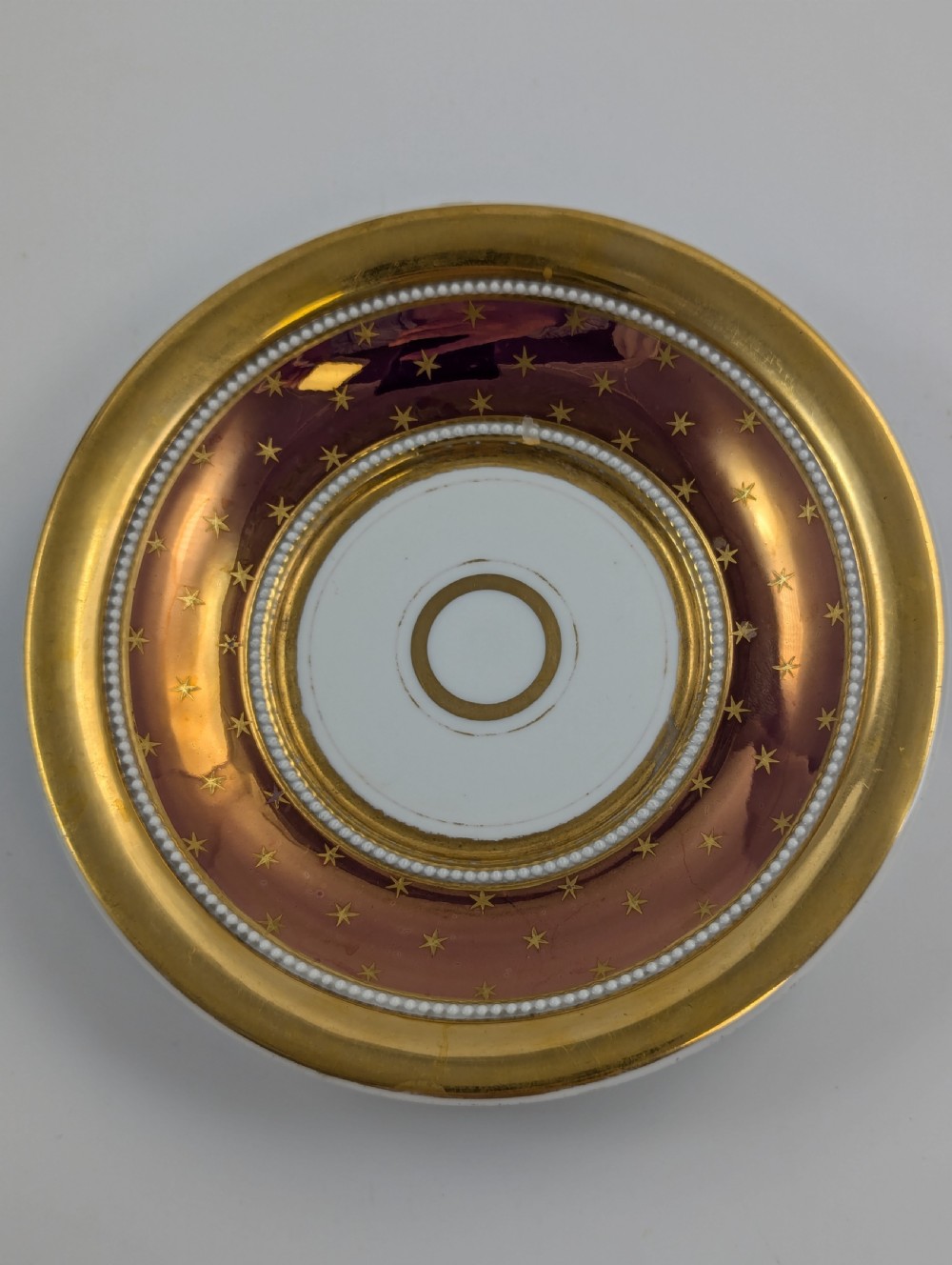 A Neoclassical KPM Berlin porcelain teacup and saucer featuring heavy gold leaf gilding, a maroon background with star patterns, and a high curved loop handle. View X