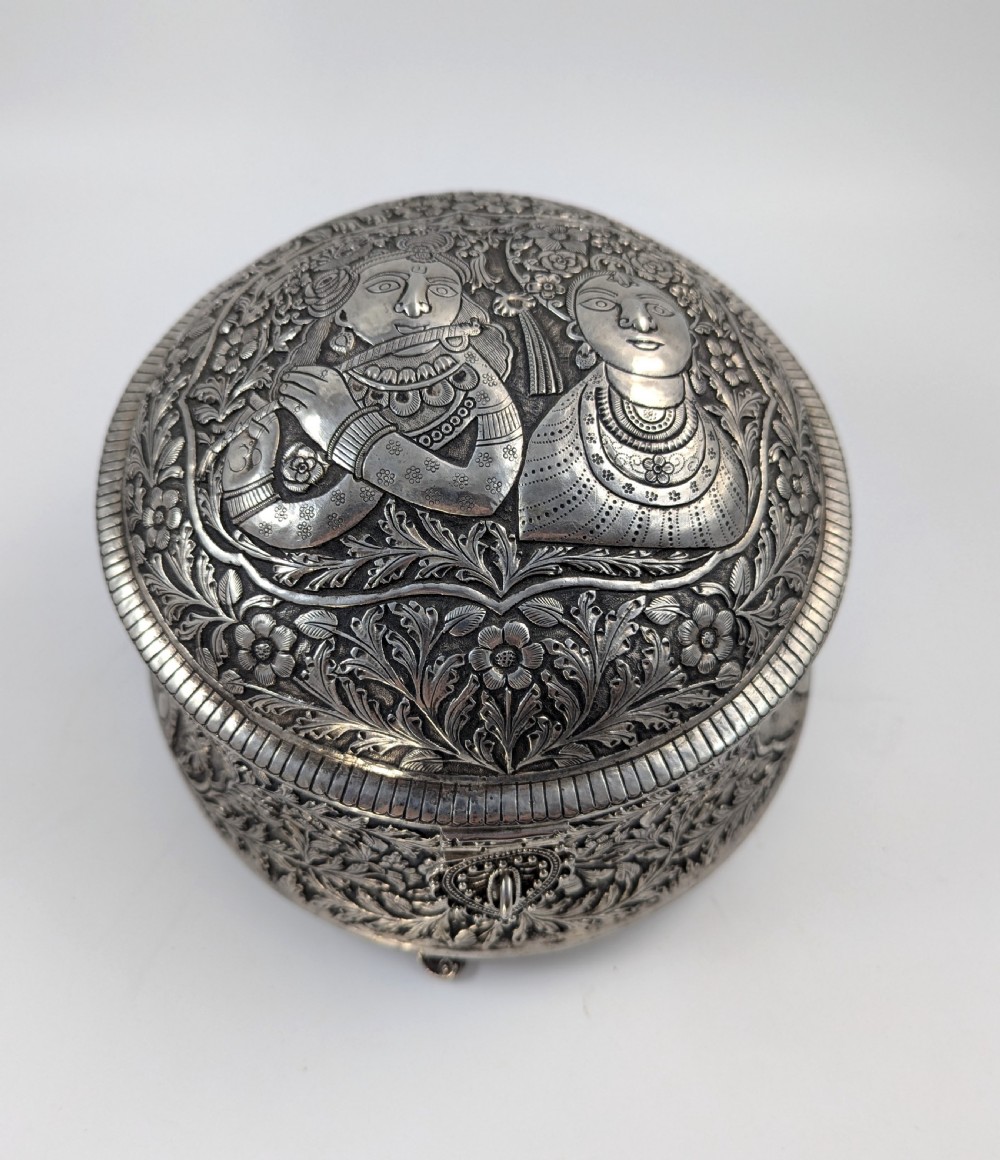 A circular silver-plated box decorated with intricate chased floral motifs and low-relief figures of Krishna and Radha on the lid.
