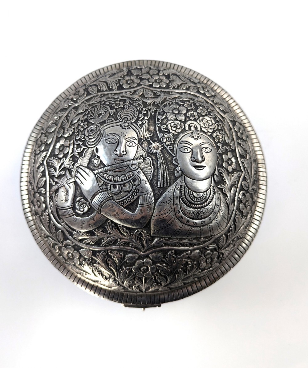 A circular silver-plated box decorated with intricate chased floral motifs and low-relief figures of Krishna and Radha on the lid. View II