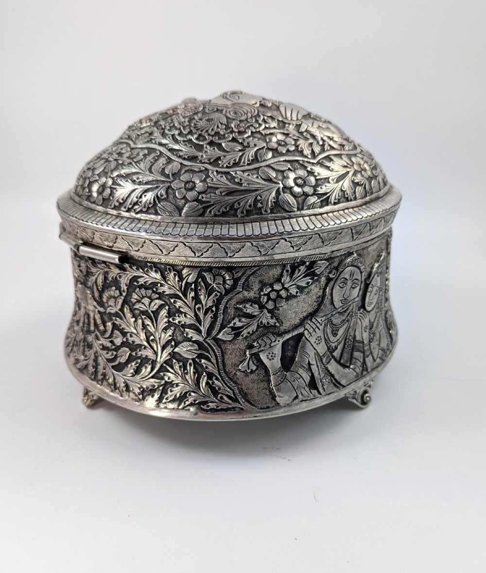 A circular silver-plated box decorated with intricate chased floral motifs and low-relief figures of Krishna and Radha on the lid. View X