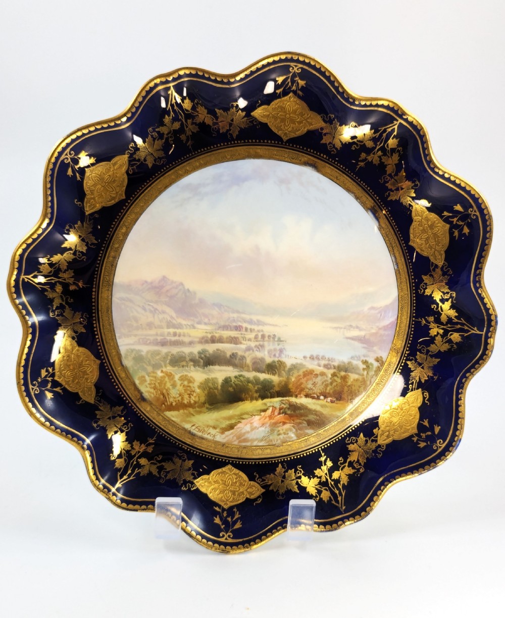 A Victorian Aynsley porcelain cabinet plate featuring a central hand-painted landscape surrounded by a scalloped cobalt blue border with intricate gold leaf ornamentation.