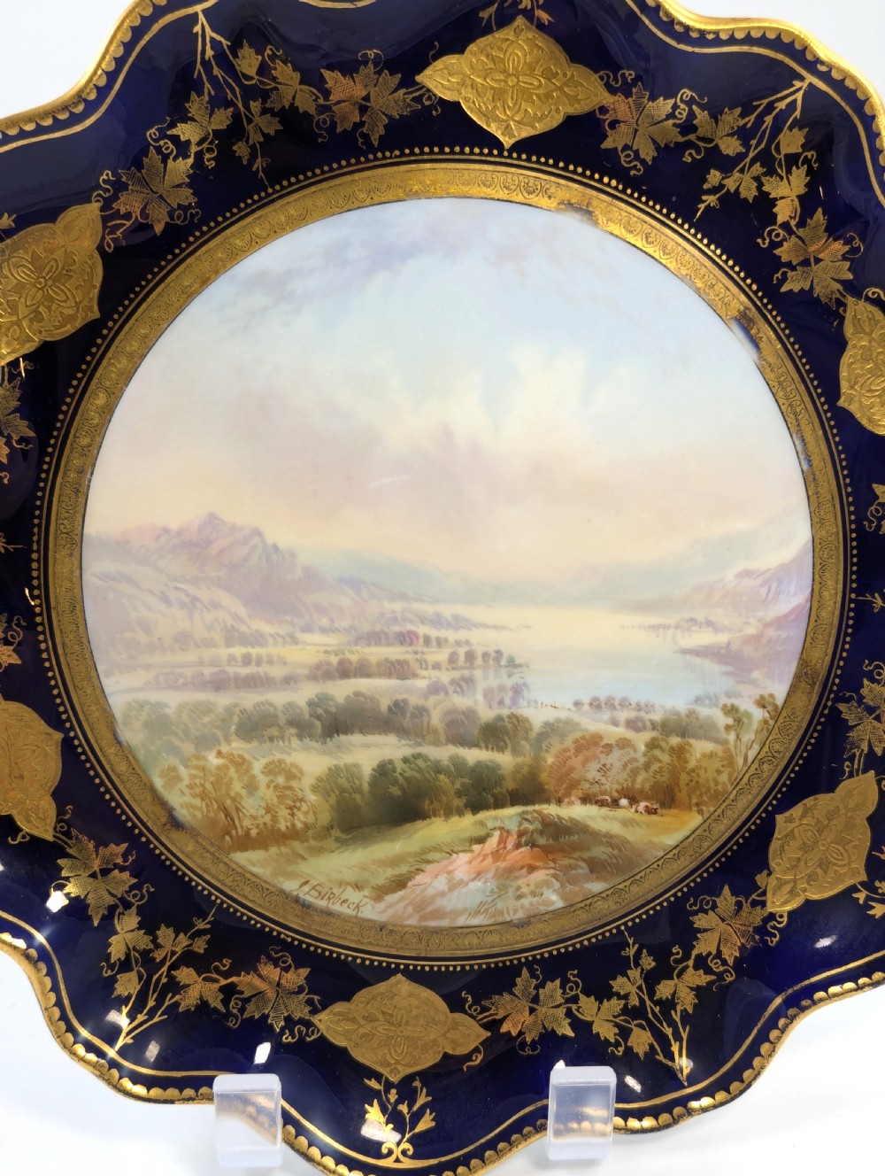 A Victorian Aynsley porcelain cabinet plate featuring a central hand-painted landscape surrounded by a scalloped cobalt blue border with intricate gold leaf ornamentation. View III