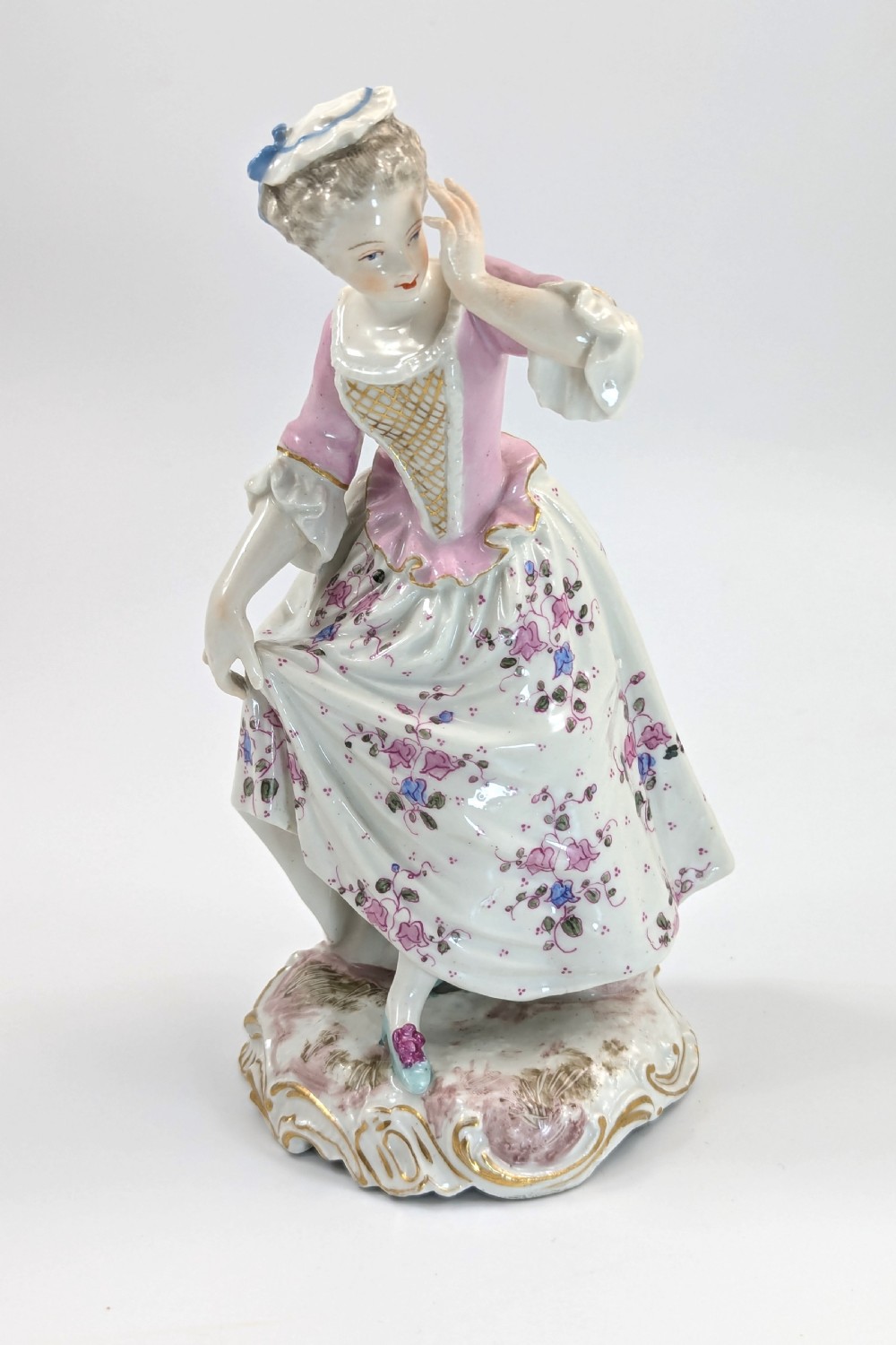 An antique French Samson porcelain figurine depicting a woman in eighteenth-century attire with a pink bodice and floral skirt standing on a scrollwork base.