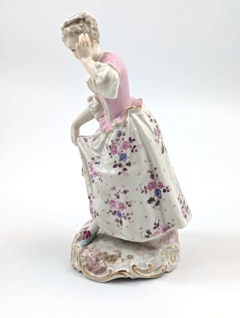An antique French Samson porcelain figurine depicting a woman in eighteenth-century attire with a pink bodice and floral skirt standing on a scrollwork base. View II