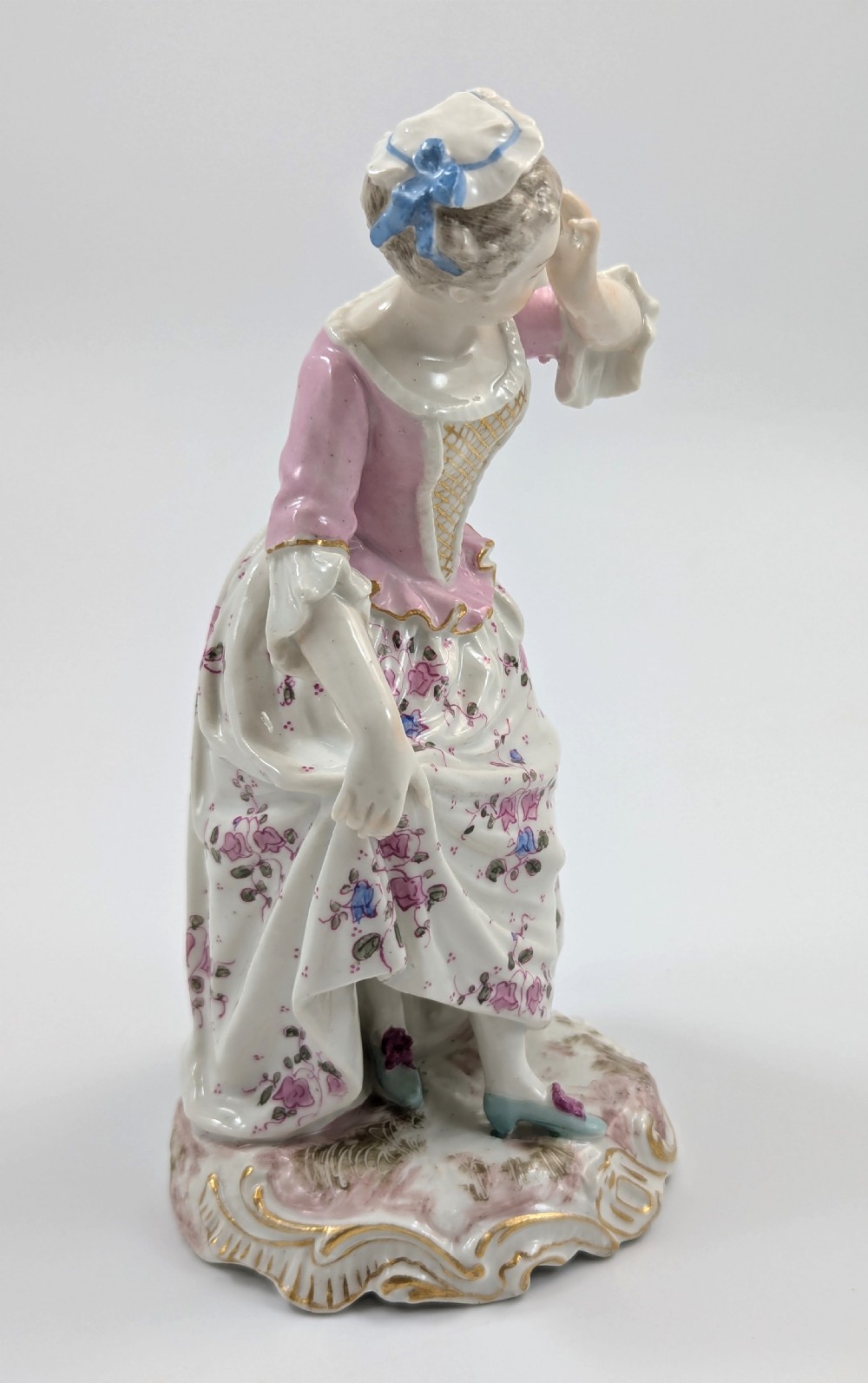 An antique French Samson porcelain figurine depicting a woman in eighteenth-century attire with a pink bodice and floral skirt standing on a scrollwork base. View IV