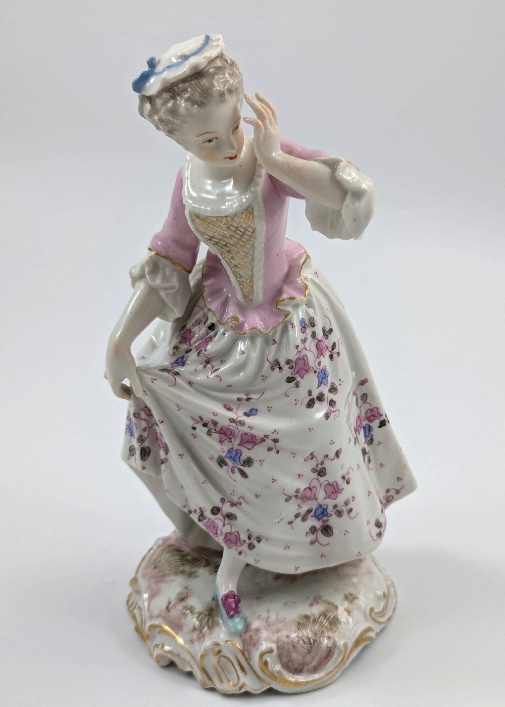 An antique French Samson porcelain figurine depicting a woman in eighteenth-century attire with a pink bodice and floral skirt standing on a scrollwork base. View VII