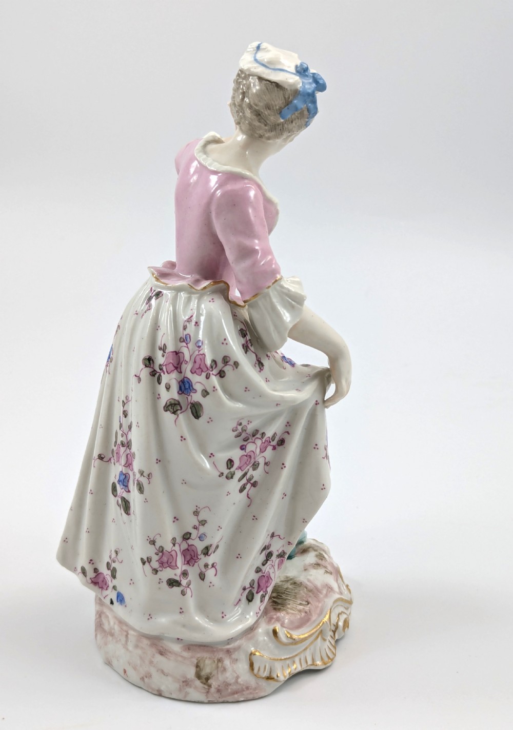 An antique French Samson porcelain figurine depicting a woman in eighteenth-century attire with a pink bodice and floral skirt standing on a scrollwork base. View X