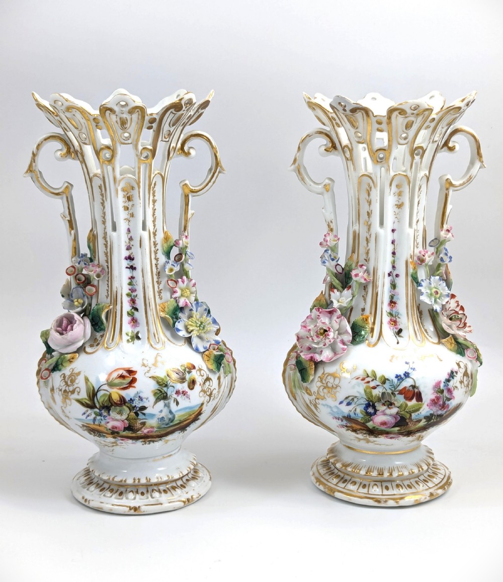 A pair of nineteenth-century white hard-paste porcelain vases featuring reticulated necks, gilded scroll handles, painted botanical panels, and applied ceramic flowers.