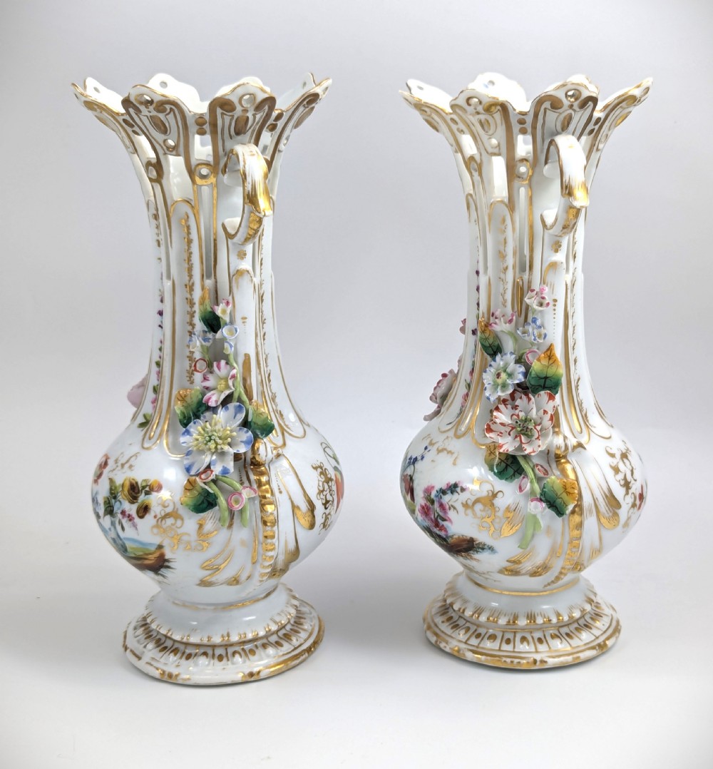 A pair of nineteenth-century white hard-paste porcelain vases featuring reticulated necks, gilded scroll handles, painted botanical panels, and applied ceramic flowers. View II