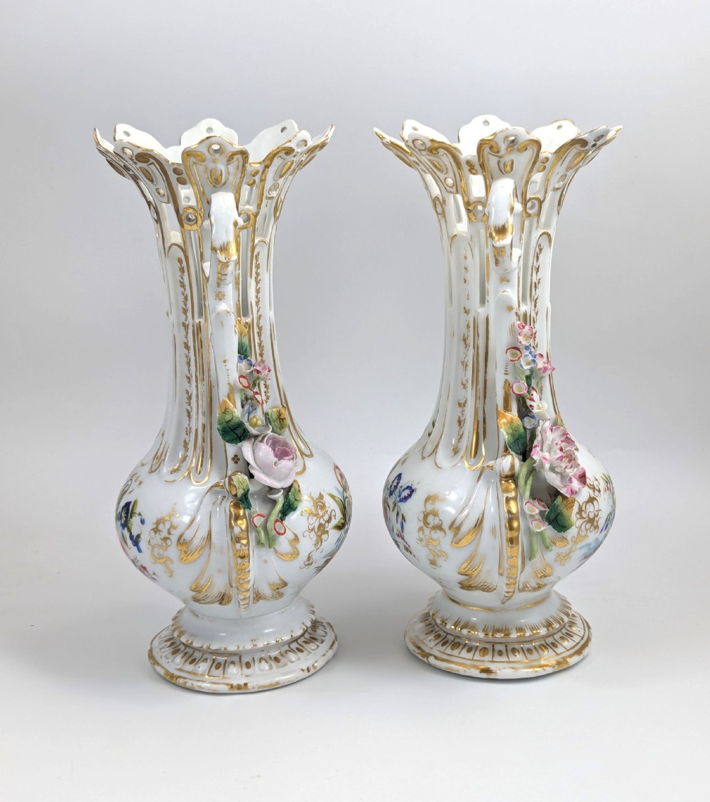 A pair of nineteenth-century white hard-paste porcelain vases featuring reticulated necks, gilded scroll handles, painted botanical panels, and applied ceramic flowers. View IV
