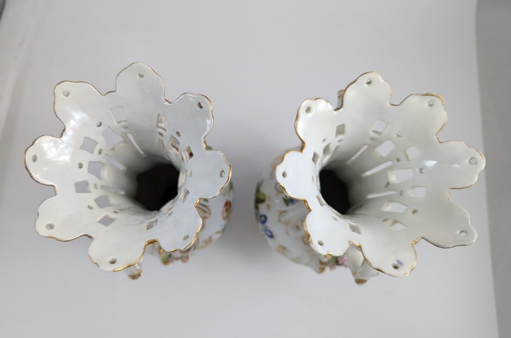 A pair of nineteenth-century white hard-paste porcelain vases featuring reticulated necks, gilded scroll handles, painted botanical panels, and applied ceramic flowers. View X