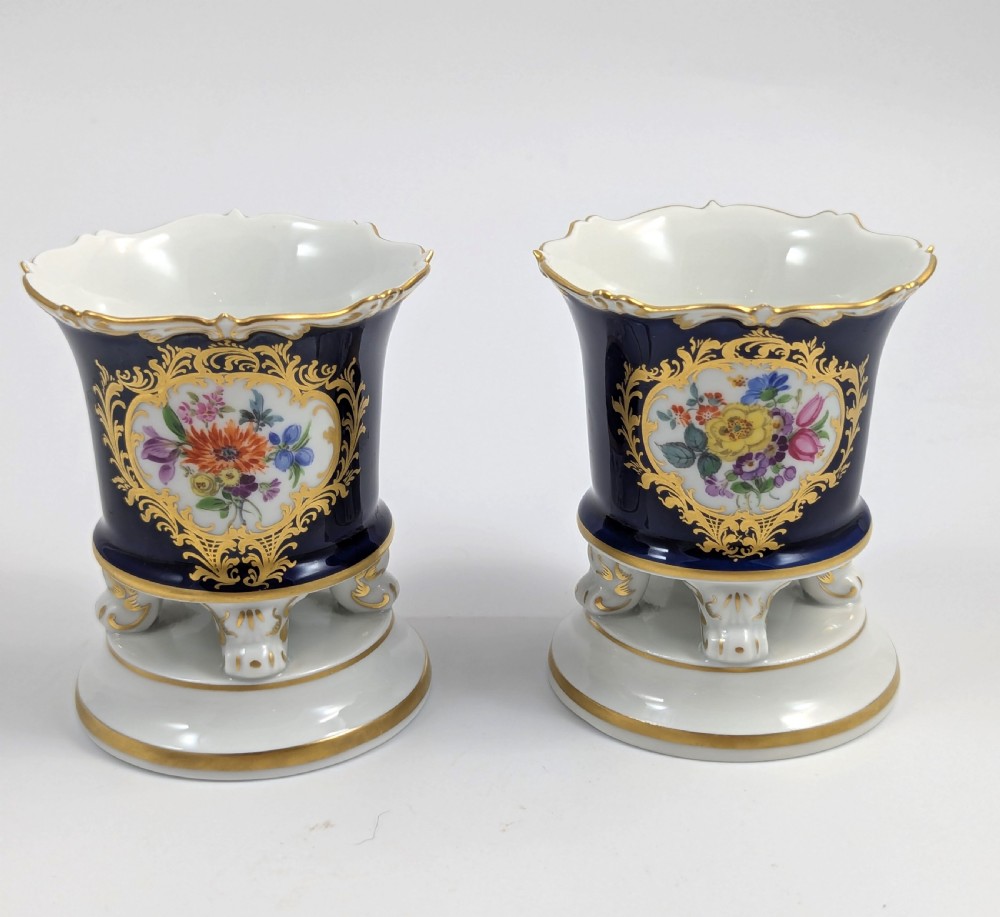 A pair of small Meissen cobalt blue porcelain vases featuring scalloped gilt rims, hand-painted floral cartouches, scroll feet, and circular white bases.