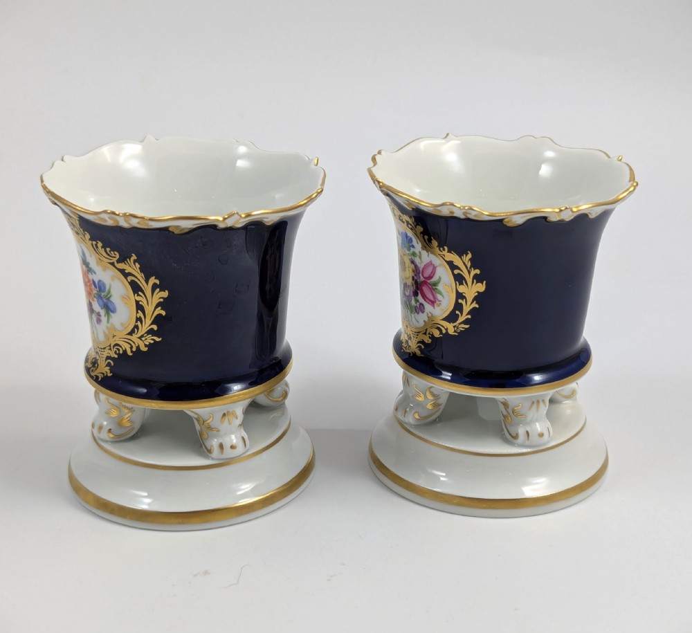 A pair of small Meissen cobalt blue porcelain vases featuring scalloped gilt rims, hand-painted floral cartouches, scroll feet, and circular white bases. View II