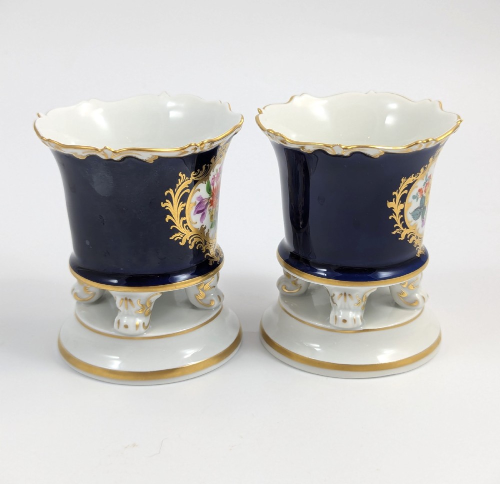 A pair of small Meissen cobalt blue porcelain vases featuring scalloped gilt rims, hand-painted floral cartouches, scroll feet, and circular white bases. View IV