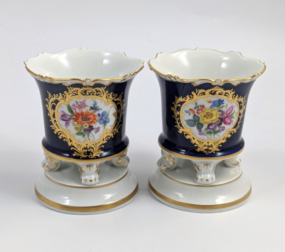 A pair of small Meissen cobalt blue porcelain vases featuring scalloped gilt rims, hand-painted floral cartouches, scroll feet, and circular white bases. View V