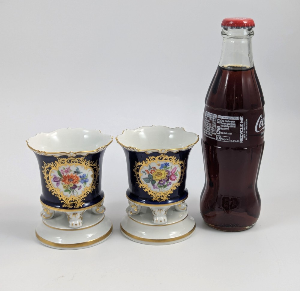 A pair of small Meissen cobalt blue porcelain vases featuring scalloped gilt rims, hand-painted floral cartouches, scroll feet, and circular white bases. View VI