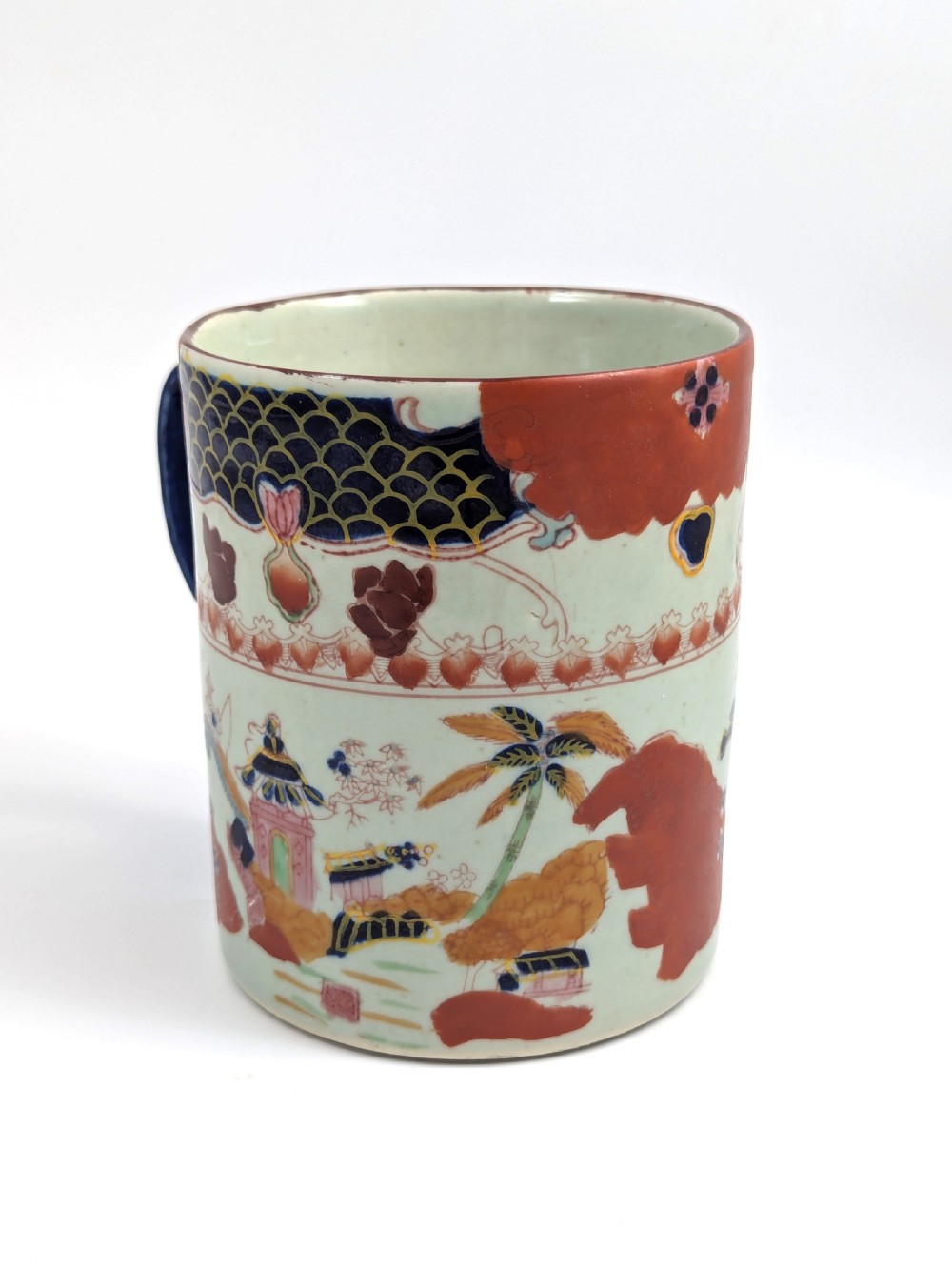 A cylindrical antique Chinese Kangxi porcelain tankard decorated with hand-painted pagodas, palm trees, and an iron-red scale pattern border. View V