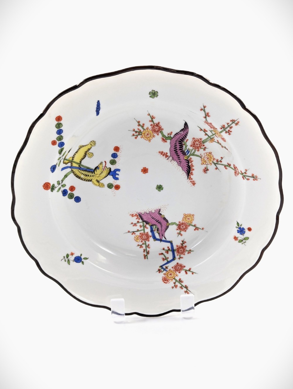 An antique white hard-paste porcelain plate decorated with hand-painted Kakiemon style motifs of a yellow tiger, purple bird, and flowering branches with a scalloped dark rim.