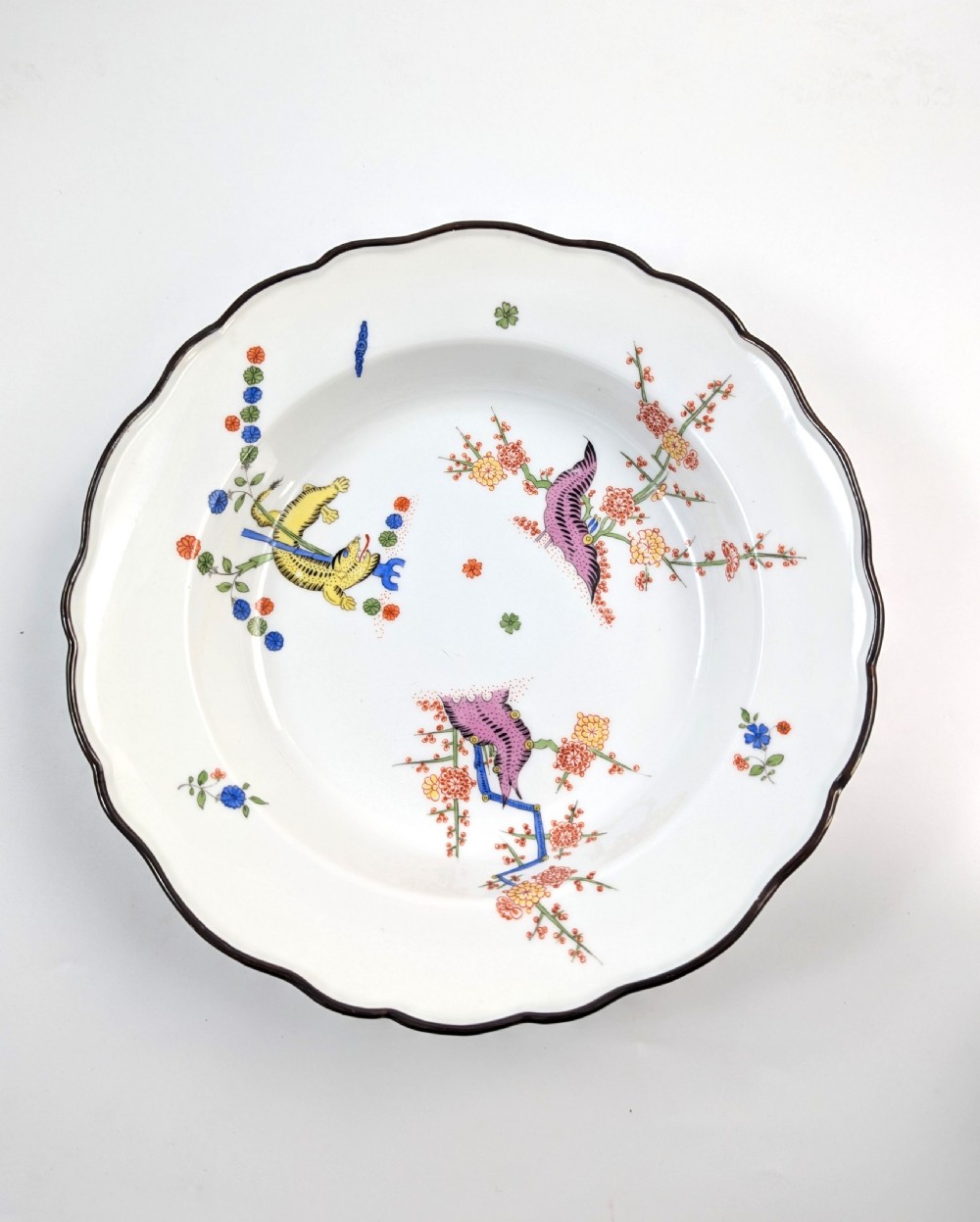 An antique white hard-paste porcelain plate decorated with hand-painted Kakiemon style motifs of a yellow tiger, purple bird, and flowering branches with a scalloped dark rim. View II