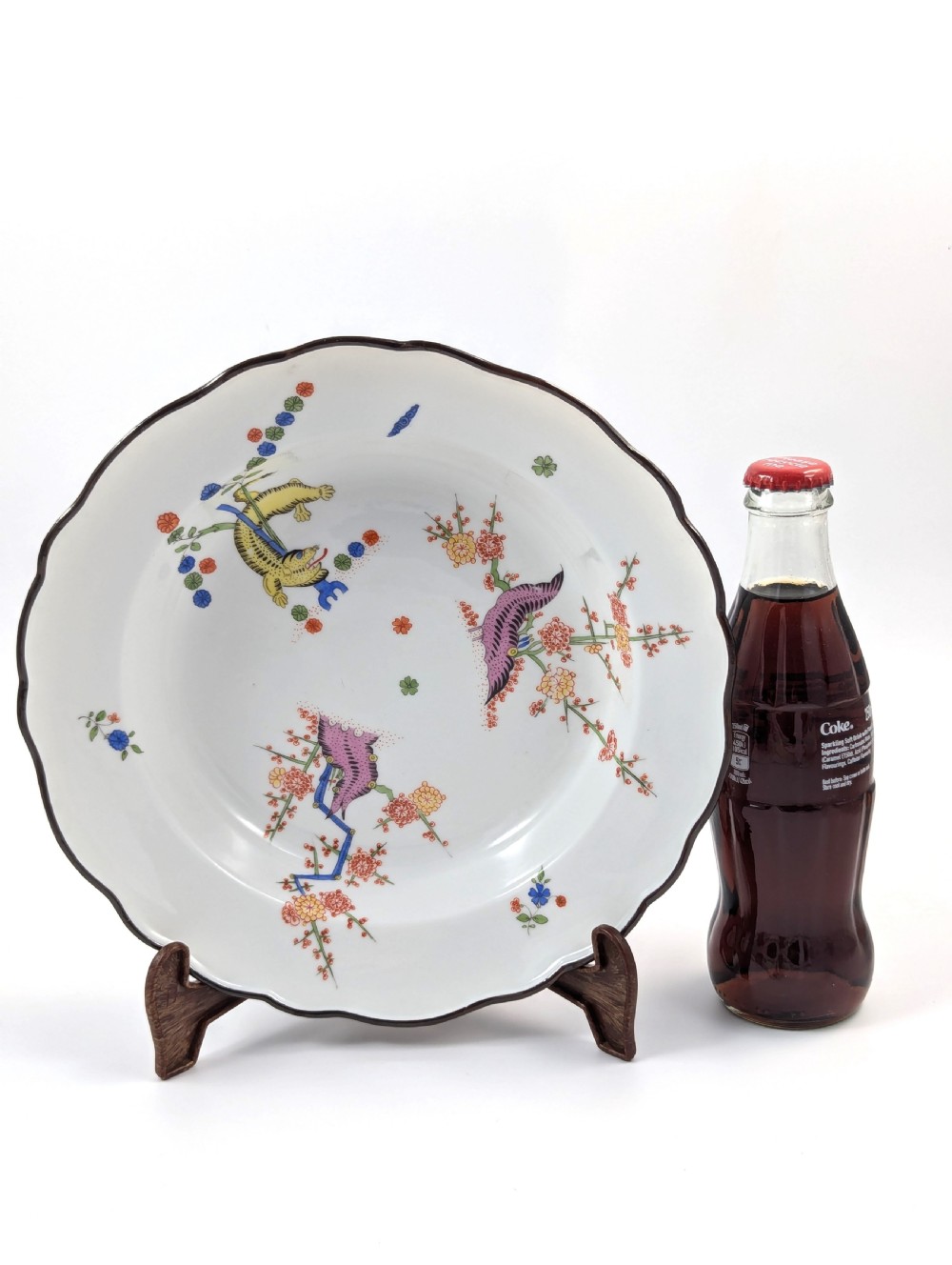 An antique white hard-paste porcelain plate decorated with hand-painted Kakiemon style motifs of a yellow tiger, purple bird, and flowering branches with a scalloped dark rim. View X
