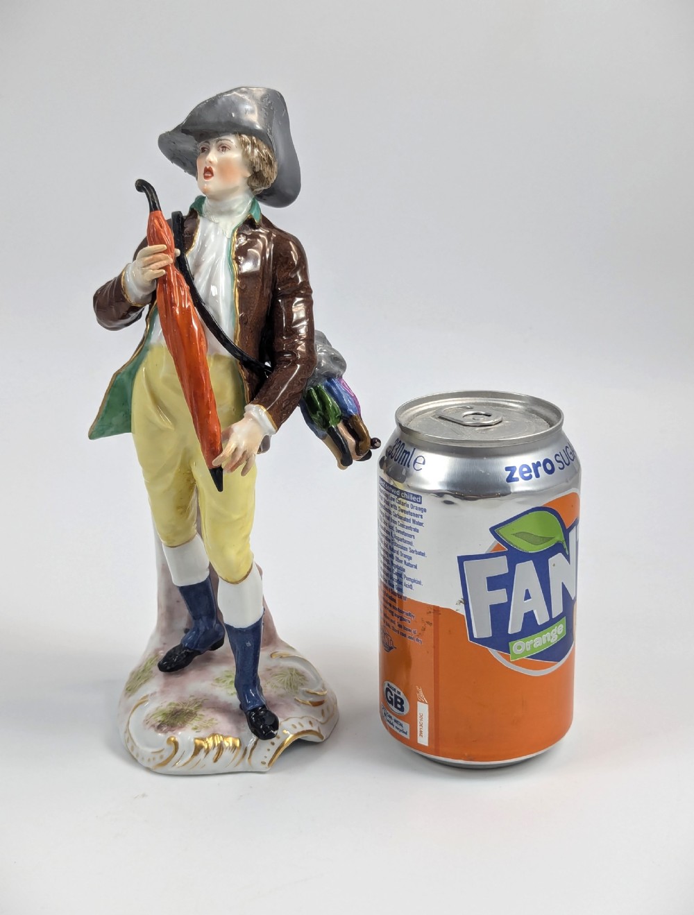 A 19th-century French Samson porcelain figurine depicting an umbrella seller in an 18th-century style tricorn hat, brown coat, and yellow breeches holding an orange umbrella. View IX