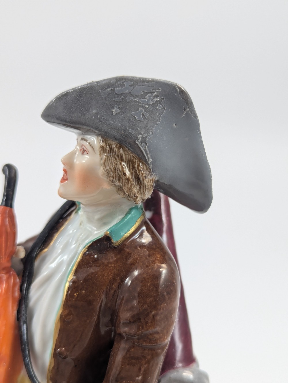 A 19th-century French Samson porcelain figurine depicting an umbrella seller in an 18th-century style tricorn hat, brown coat, and yellow breeches holding an orange umbrella. View X