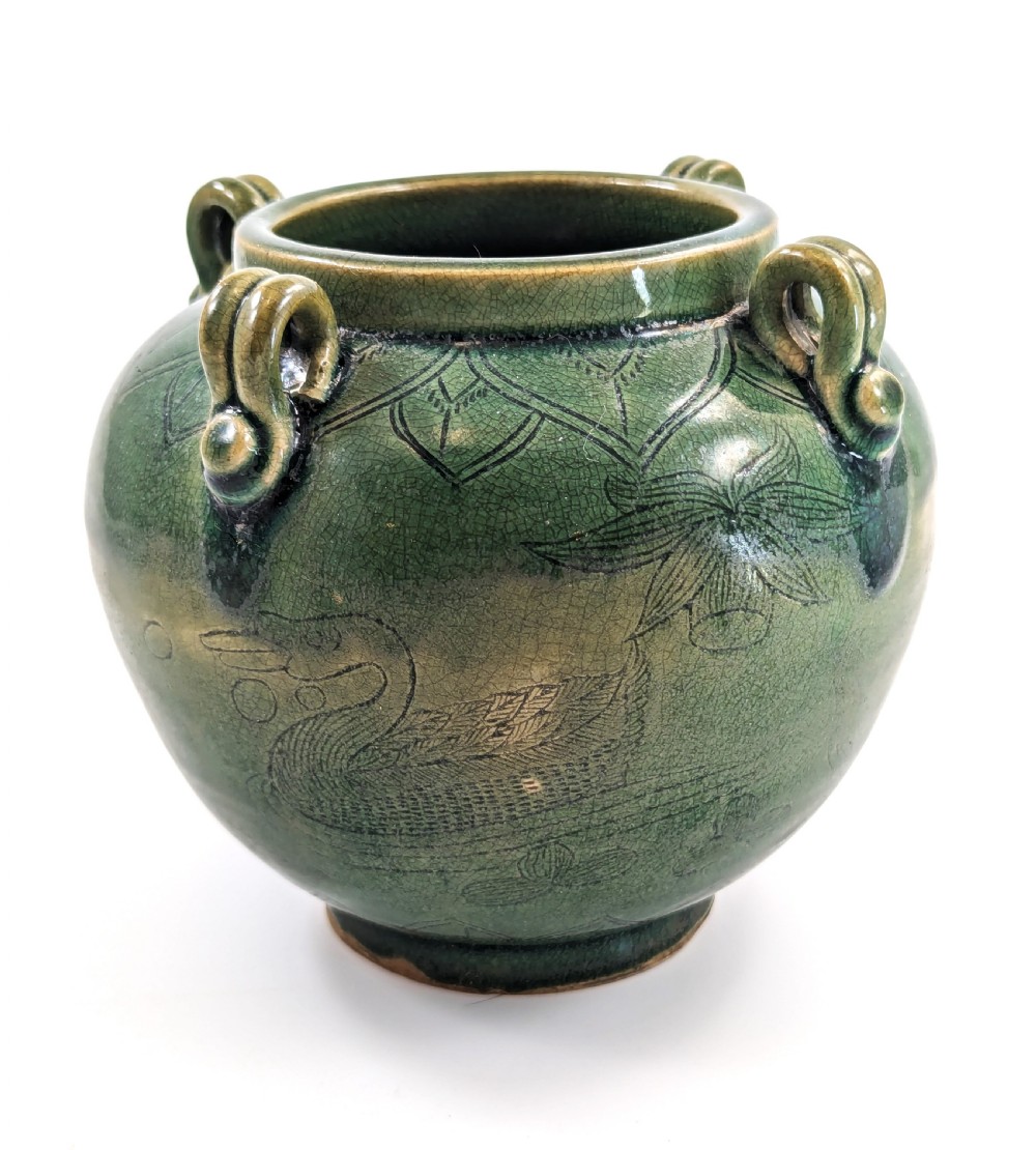 A rounded olive green celadon ceramic jar with four loop handles along the rim, featuring crackle glaze and incised line drawings of ducks and lotus plants.