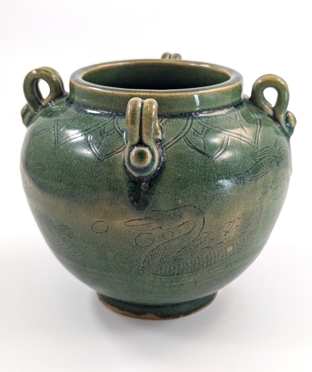 A rounded olive green celadon ceramic jar with four loop handles along the rim, featuring crackle glaze and incised line drawings of ducks and lotus plants. View VI