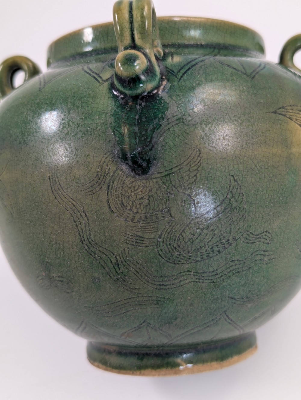 A rounded olive green celadon ceramic jar with four loop handles along the rim, featuring crackle glaze and incised line drawings of ducks and lotus plants. View XI