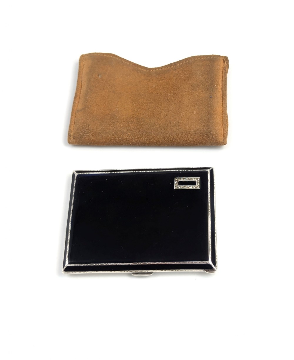 An Art Deco rectangular sterling silver cigarette case featuring a polished black enamel cover with a marcasite-inset rectangular corner detail and a beige suede pouch.