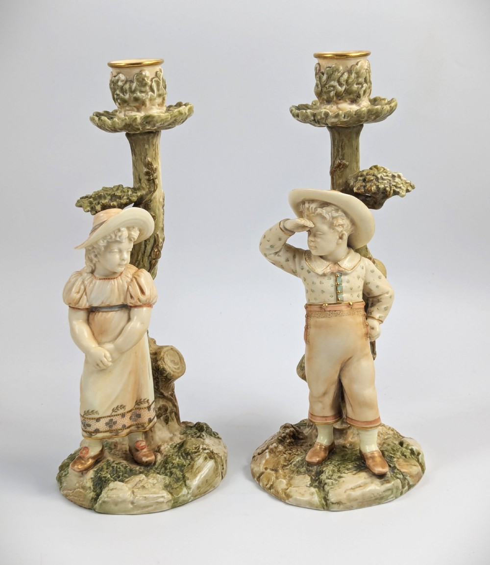 A pair of Victorian Royal Worcester porcelain candlesticks featuring a young boy and girl standing before tree trunk columns topped with leafy candle sconces.