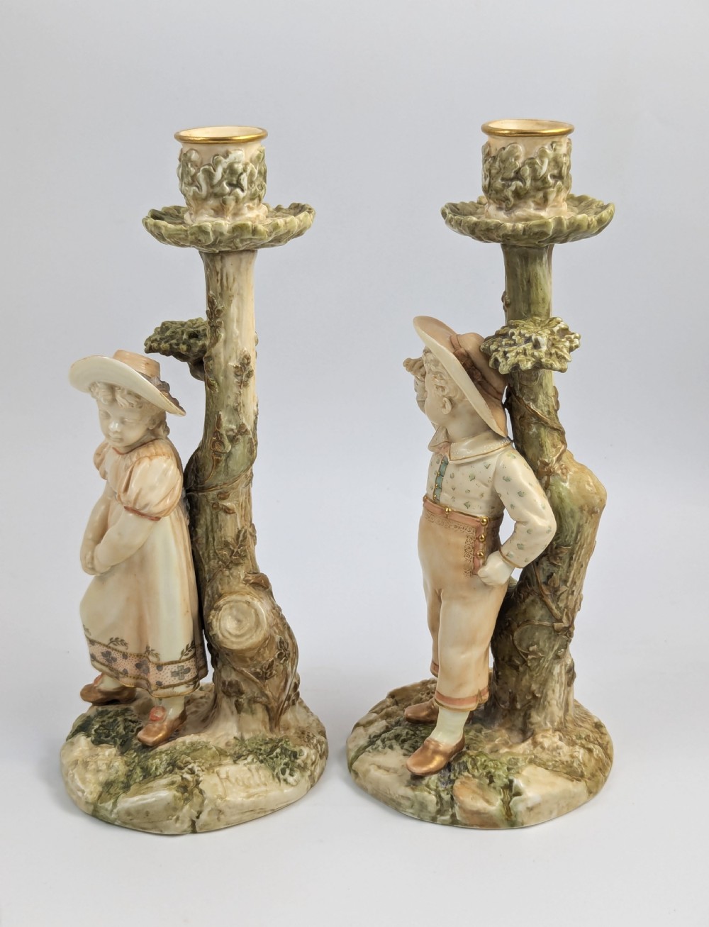 A pair of Victorian Royal Worcester porcelain candlesticks featuring a young boy and girl standing before tree trunk columns topped with leafy candle sconces. View II