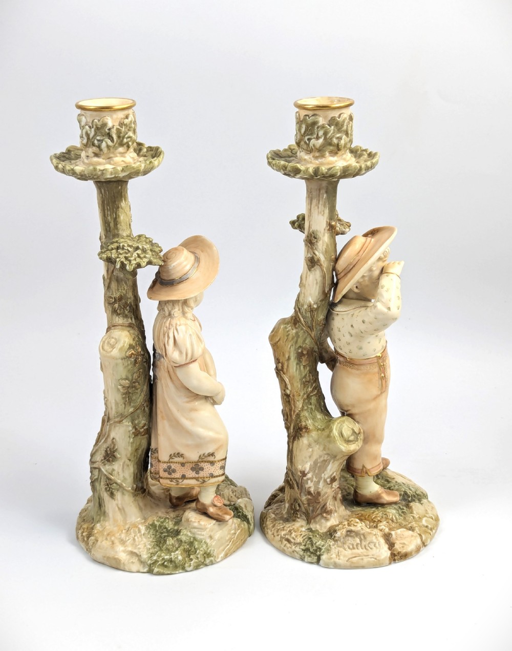 A pair of Victorian Royal Worcester porcelain candlesticks featuring a young boy and girl standing before tree trunk columns topped with leafy candle sconces. View V