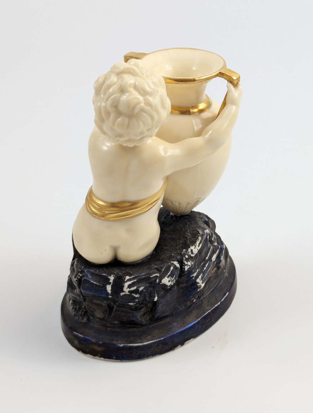 An ivory-coloured porcelain figurine depicting a small child seated beside a gilded amphora vessel on a dark base. View II
