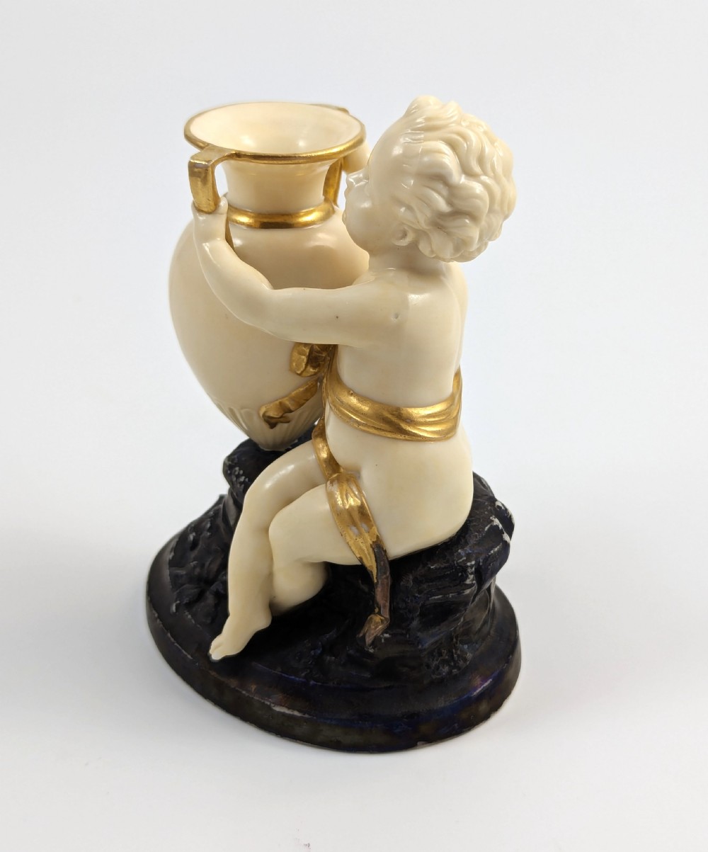 An ivory-coloured porcelain figurine depicting a small child seated beside a gilded amphora vessel on a dark base. View III