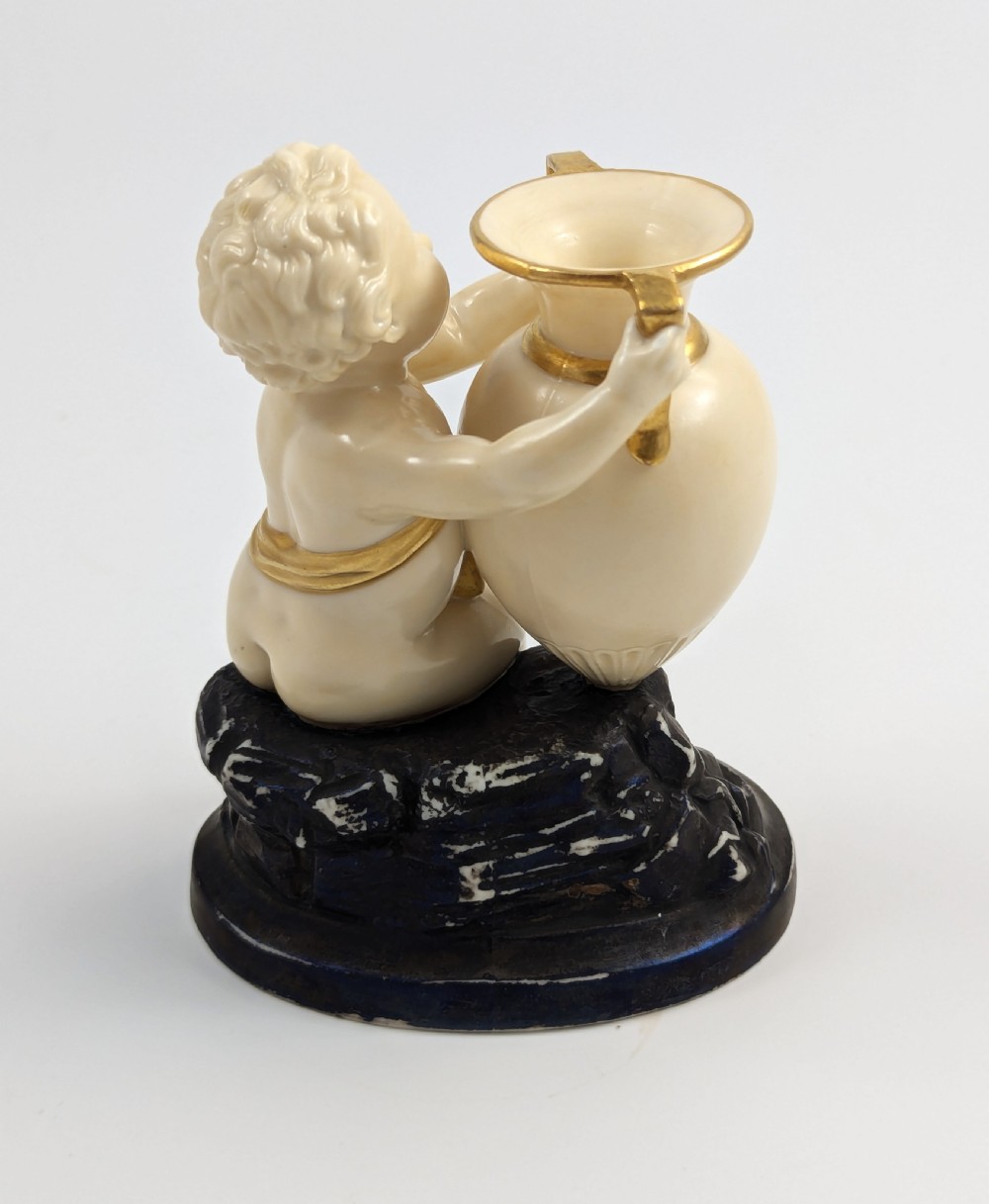 An ivory-coloured porcelain figurine depicting a small child seated beside a gilded amphora vessel on a dark base. View IV