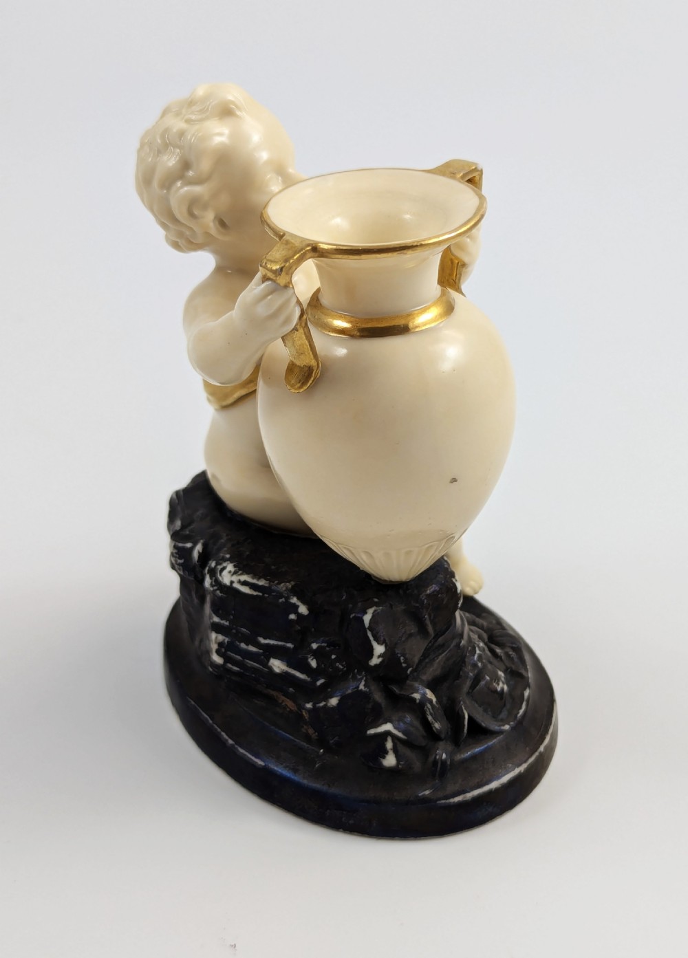 An ivory-coloured porcelain figurine depicting a small child seated beside a gilded amphora vessel on a dark base. View V