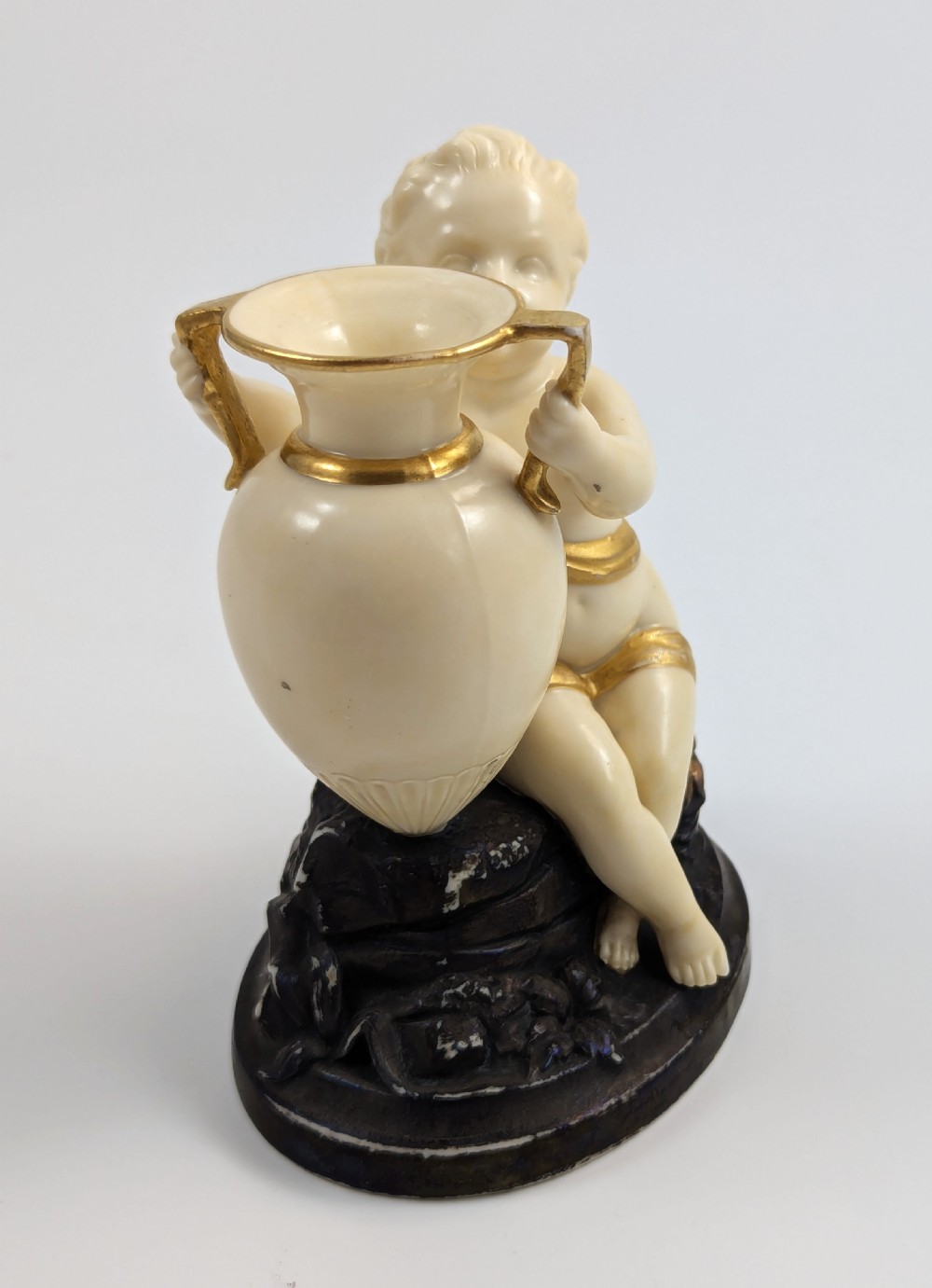 An ivory-coloured porcelain figurine depicting a small child seated beside a gilded amphora vessel on a dark base. View VI