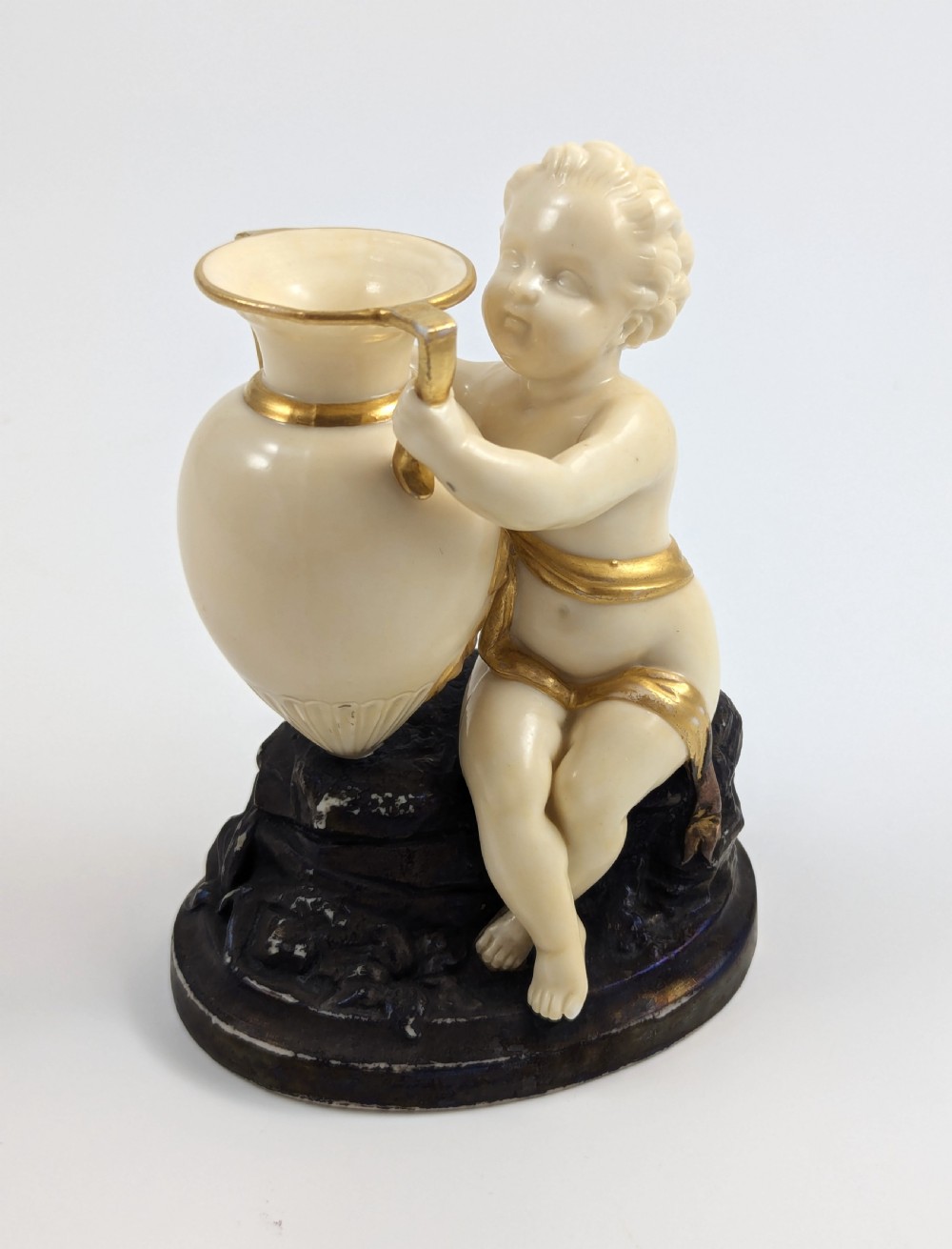 An ivory-coloured porcelain figurine depicting a small child seated beside a gilded amphora vessel on a dark base. View VII