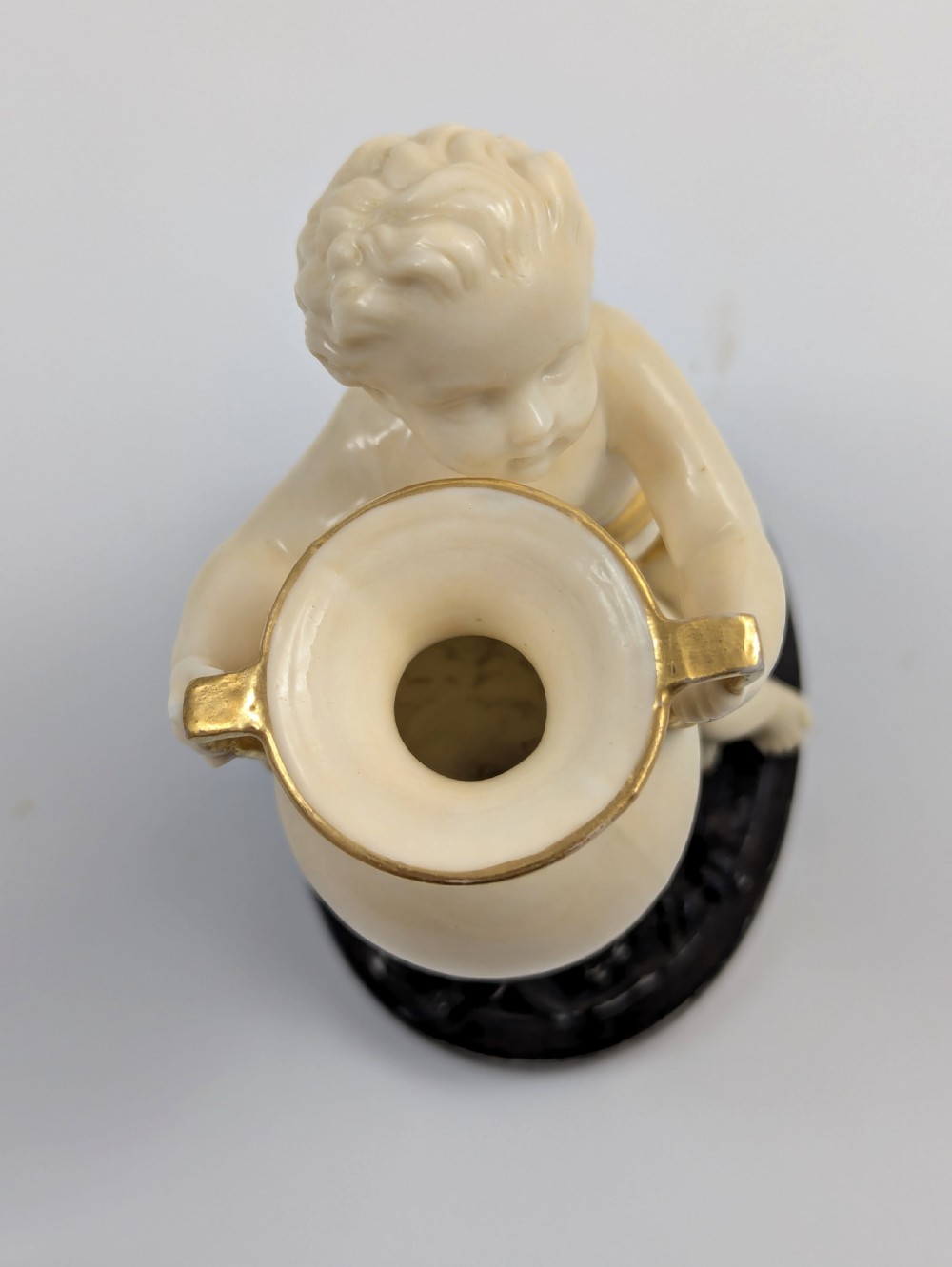 An ivory-coloured porcelain figurine depicting a small child seated beside a gilded amphora vessel on a dark base. View VIII